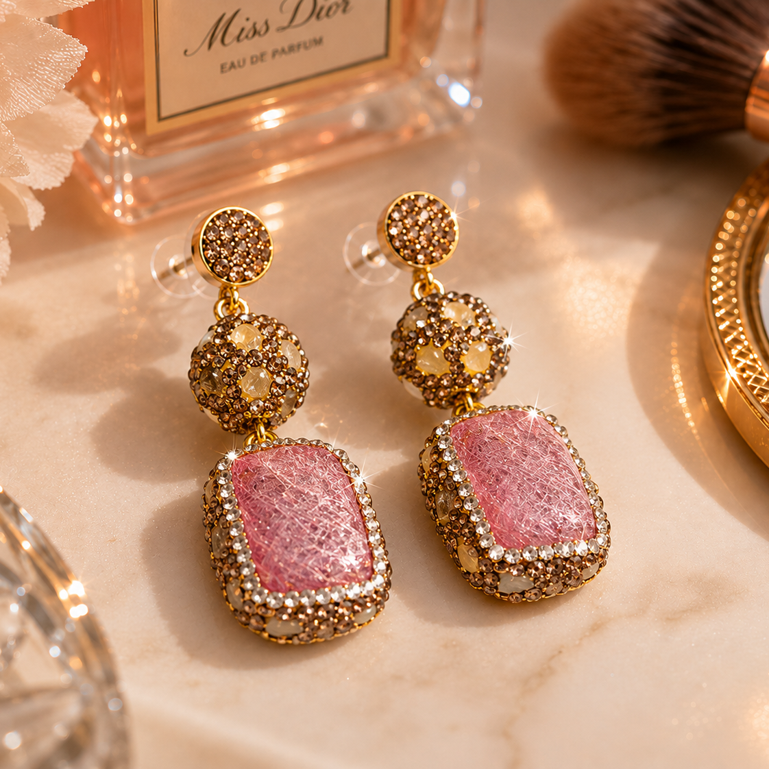 A pair of gold-toned statement dangle earrings with rectangular pink glitter stones, displayed on a marble surface with perfume, makeup brushes, and a compact mirror in the background.